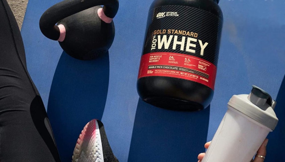 a large container of whey protein powder resting on a yoga mat with a weight on it next to a person holding a protein shake 