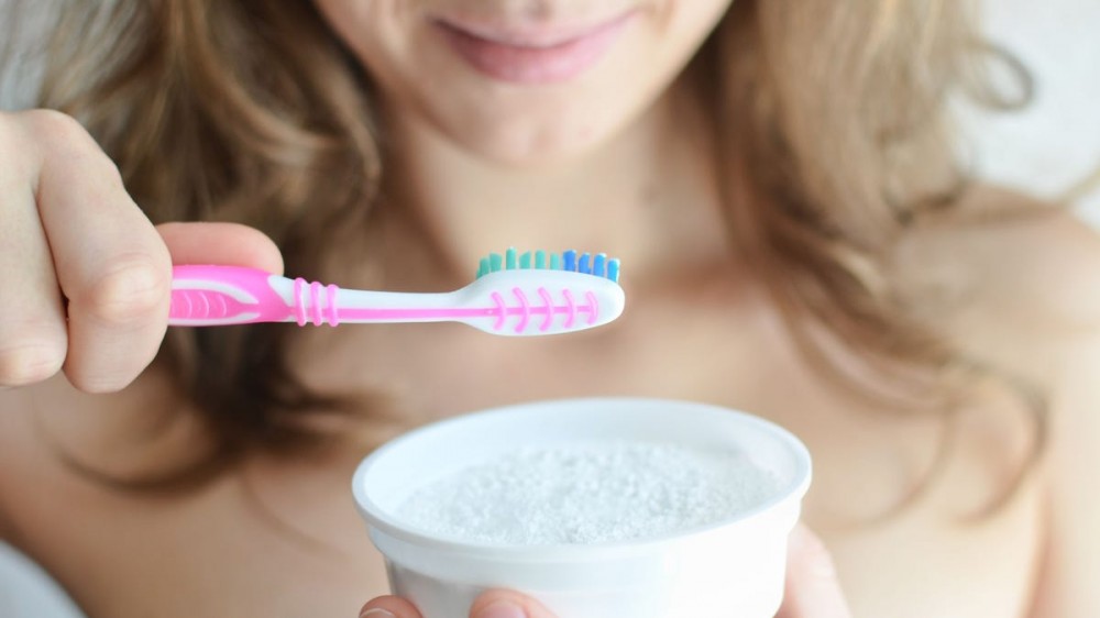 A woman holds up a toothbrush and a cup of baking soda.