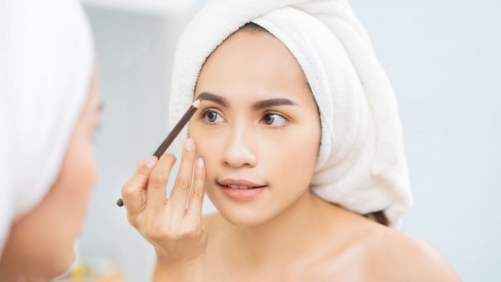 A woman applies eyebrow pencil to her eyebrows while looking in the mirror.