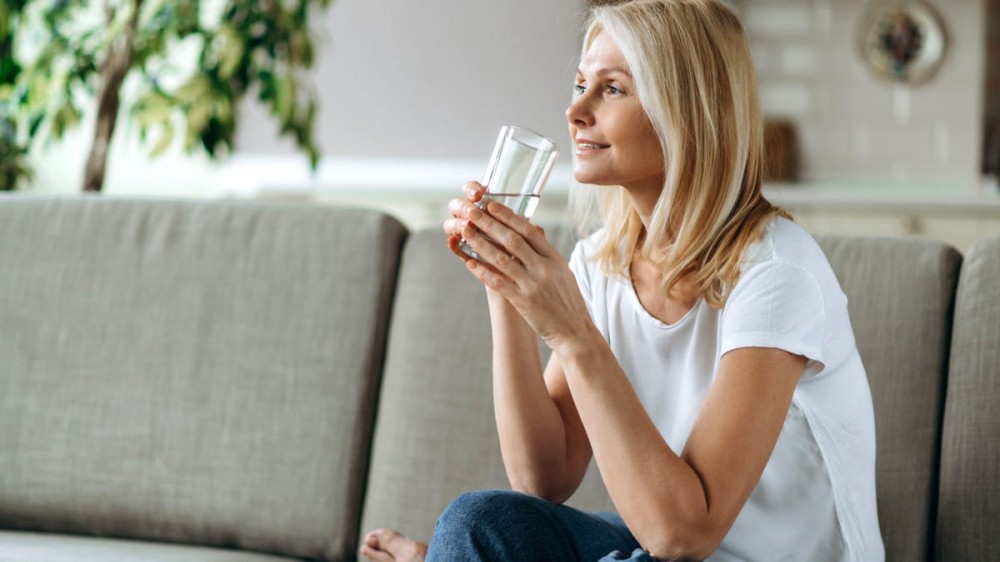 A blonde woman sits on the couch and drinks a tall glass of water.