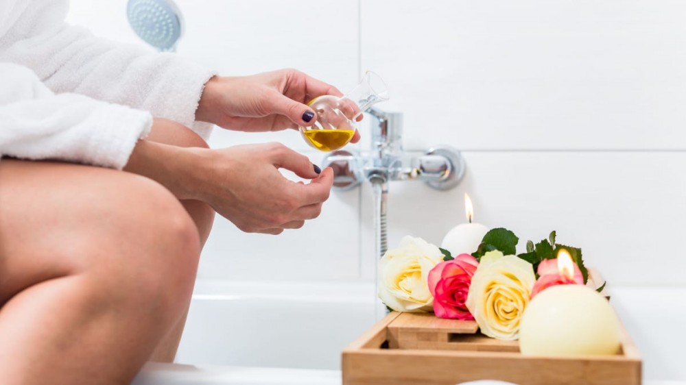 Woman preparing wellness bath with flowers, candles, and fragrance oil in tub.