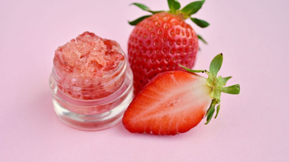 Two strawberries sit next to a glass jar of lip scrub.