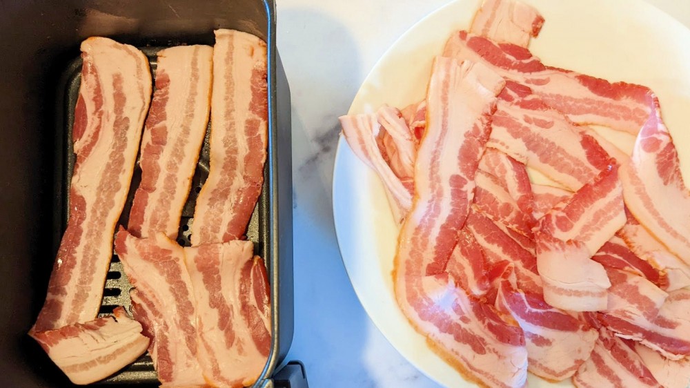 An air fryer basket with a few slices of bacon and a plate full of bacon slices.