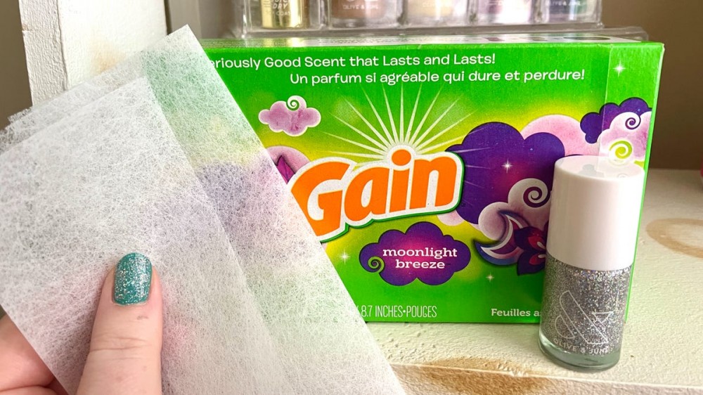 A person holds up a dryer sheet in front of a pack of dryer sheets sitting next to a bottle of glitter nail polish.