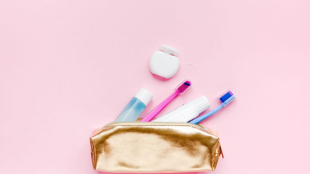 Teeth hygiene and oral dental care products in golden travel cosmetic purse kit pastel pink color background with copy space. Blank tube of toothpaste and toothbrushes. Flat lay, top view composition.