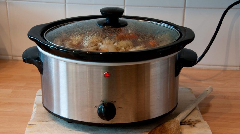 A slow cooker sits on a wooden countertop with a wooden spoon next to it.