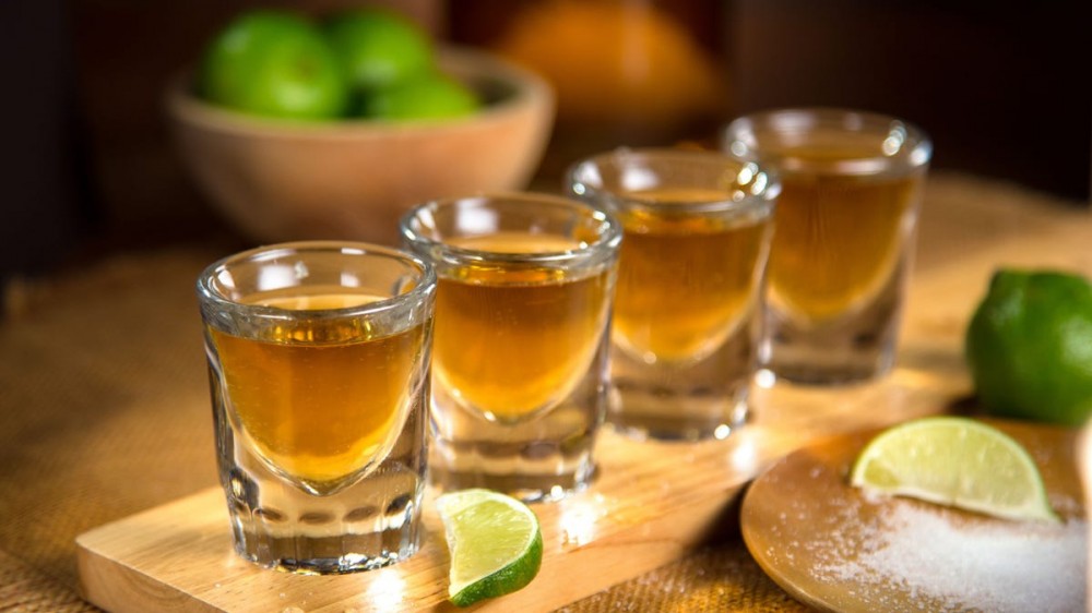 Four tequila shots sit on a wooden board with limes around them.