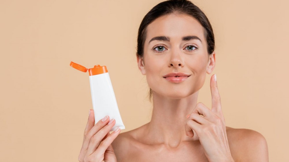 A woman holds up a tube of sunscreen while applying it to her face.