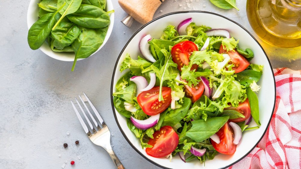 A salad with mixed green and tomatoes sits next to a fork and a bowl of fresh spinach.