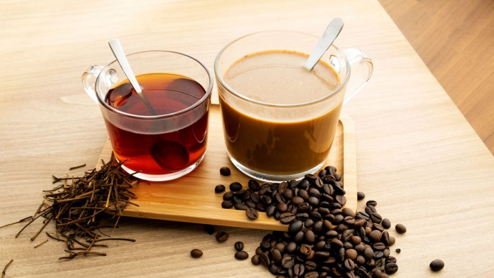 A cup of tea and a cup of coffee sit on a wooden cutting board surrounded by tea leaves and coffee beans.