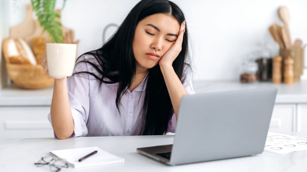 A woman falls asleep while holding a cup of coffee in front of a computer.