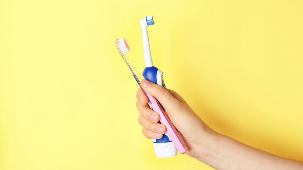 A person holds a manual toothbrush and an electric toothbrush