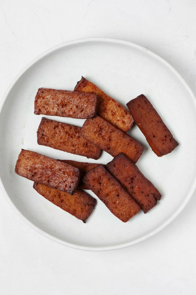 A round, white plate has been covered with rectangular, dark brown slices of baked, smoky tofu.