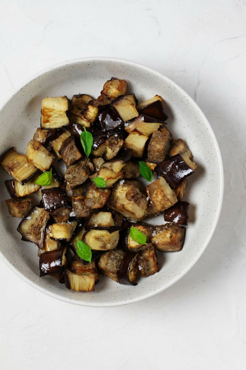 A white, ceramic bowl is filled with roasted vegetables and tiny leaves of fresh, green basil. 