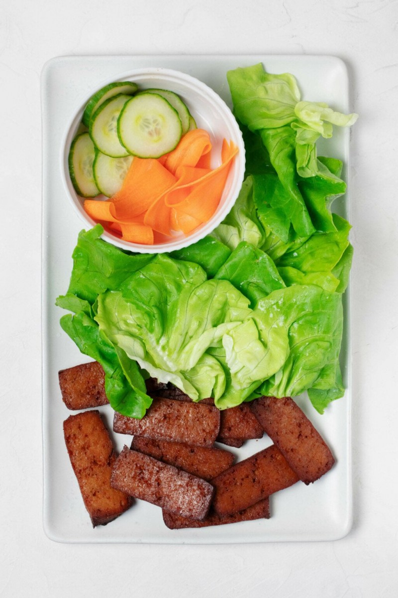 A white tray, which rests on a white surface, holds components for making sandwiches. This includes smoky tofu slices, bright green lettuce leaves, and pickled cucumbers and carrots.