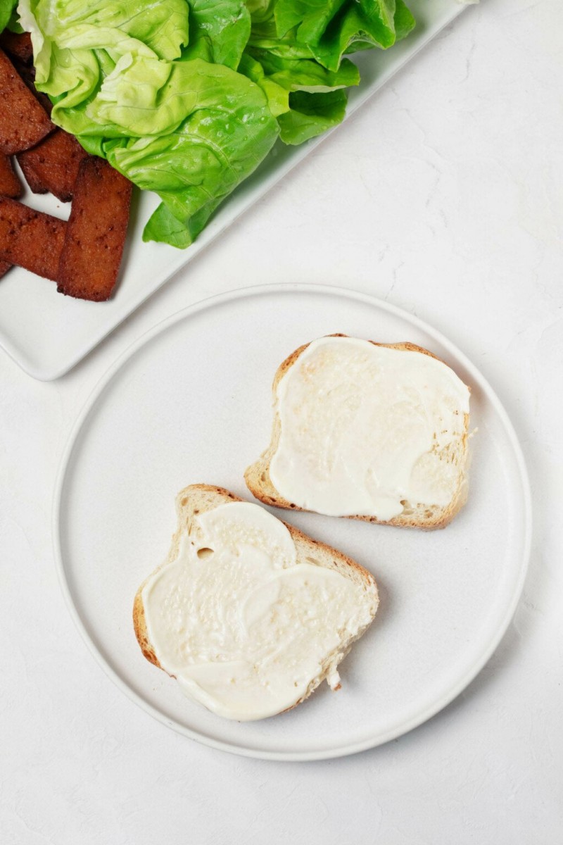 An overhead image of the setup for preparing a sandwich. Two slices of bread are coated with creamy, cream coloed mayonnaise. A tray with vegetables and tofu is nearby.