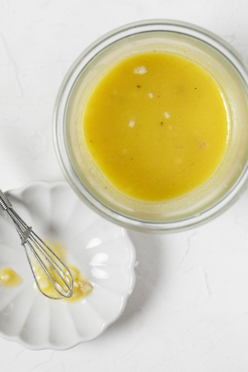 An overhead image of a pale yellow, vegan Champagne vinaigrette, with a tiny whisk resting nearby.