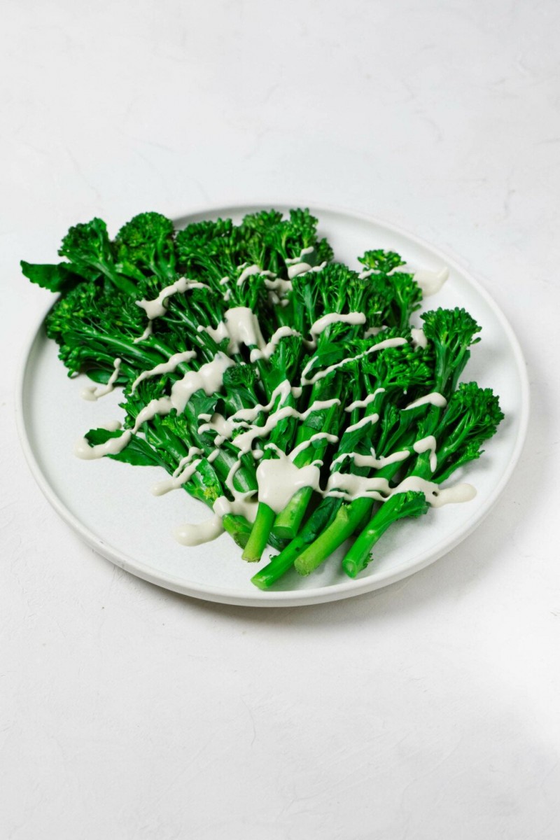 An overhead image of bright green, steamed broccolini resting on a white, round plate. The broccolini is drizzled with a creamy white tahini dressing.