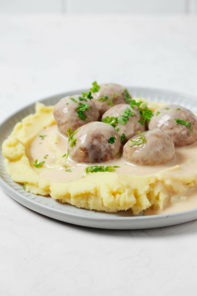 A round, gray white plate is topped with mashed potatoes, vegan Swedish meatballs, and chopped parsley.