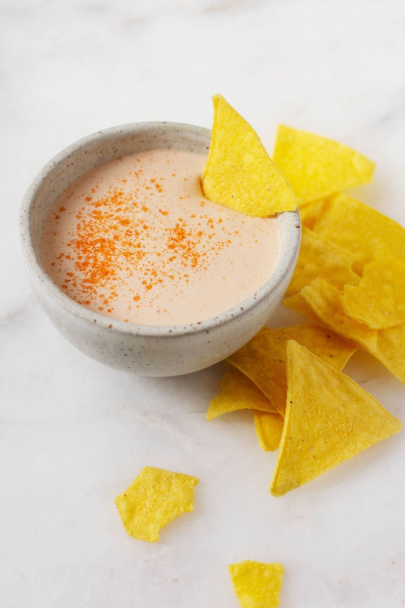 A small, circular bowl of a creamy vegan nacho sauce is accompanied by corn chips. One of the corn chips has been dipped into the sauce.